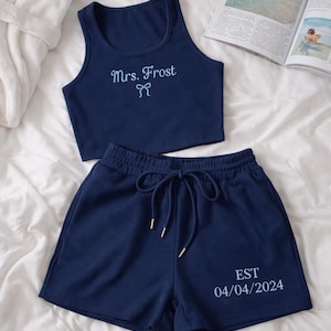 May include: Navy blue two-piece pajama set. The crop top is embroidered with "Mrs. Frost" and a bow. The shorts have a drawstring and the text "EST 04/04/2024". The set is made of a ribbed fabric.