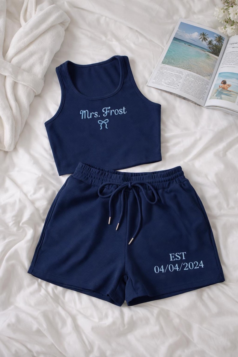 May include: Navy blue two-piece pajama set. The crop top is embroidered with "Mrs. Frost" and a bow. The shorts have a drawstring and the text "EST 04/04/2024". The set is made of a ribbed fabric.