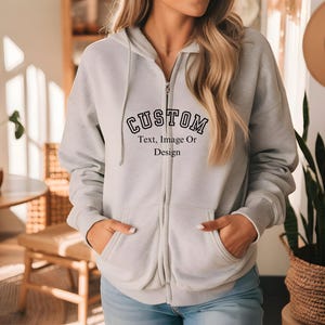 Puede incluir: Una sudadera con capucha gris claro con cremallera y la palabra "CUSTOM" en letras negras en la parte delantera. Debajo, el texto dice "Text, Image Or Design". La sudadera tiene una capucha con cordón y dos bolsillos delanteros.