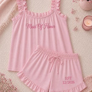 May include: A light pink pajama set featuring a ruffled tank top with the embroidered text "Maid Of Honor" and matching shorts. The shorts have a drawstring waist and the embroidered text "EST 12/2026".