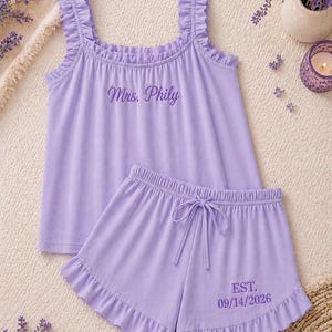 May include: A lavender pajama set featuring a tank top with ruffle straps and the embroidered text "Mrs. Phily". The matching shorts have a drawstring waist and the embroidered text "EST. 09/14/2026".