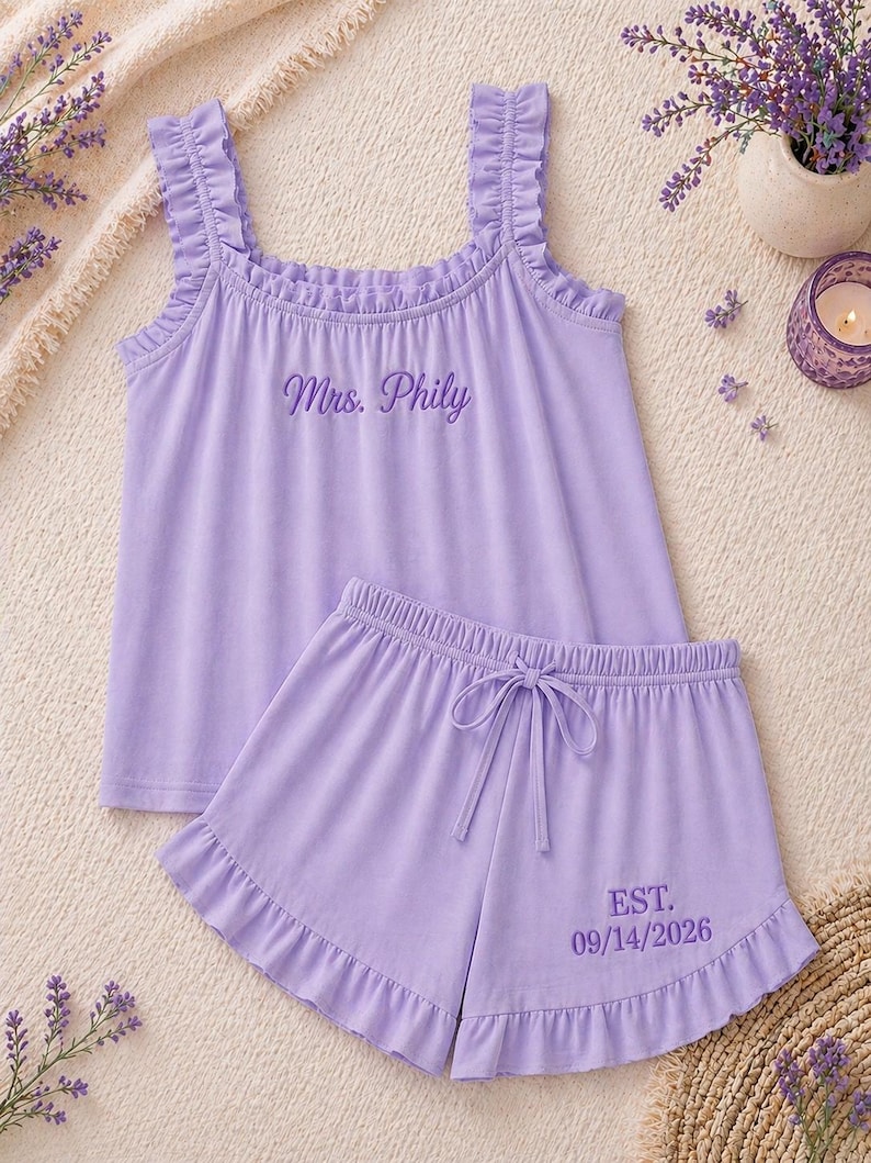 May include: A lavender pajama set featuring a tank top with ruffle straps and the embroidered text "Mrs. Phily". The matching shorts have a drawstring waist and the embroidered text "EST. 09/14/2026".