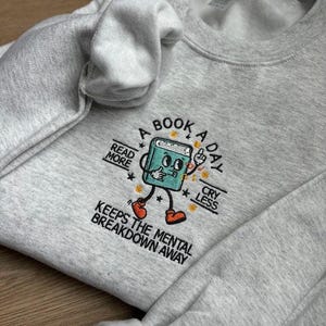 K&ouml;nnte beinhalten: Ein graues Sweatshirt mit einem gestickten Design eines Buches mit Beinen und Armen. Das Buch tr&auml;gt rote Schuhe und hat ein fr&ouml;hliches Gesicht. Der Text auf dem Sweatshirt lautet "A Book A Day Keeps The Mental Breakdown Away". Das Buch ist von Sternen umgeben und der Text "Read More" und "Cry Less" befindet sich auf beiden Seiten.