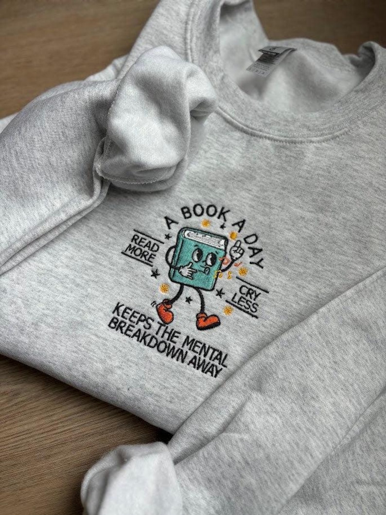 K&ouml;nnte beinhalten: Ein graues Sweatshirt mit einem gestickten Design eines Buches mit Beinen und Armen. Das Buch tr&auml;gt rote Schuhe und hat ein fr&ouml;hliches Gesicht. Der Text auf dem Sweatshirt lautet "A Book A Day Keeps The Mental Breakdown Away". Das Buch ist von Sternen umgeben und der Text "Read More" und "Cry Less" befindet sich auf beiden Seiten.