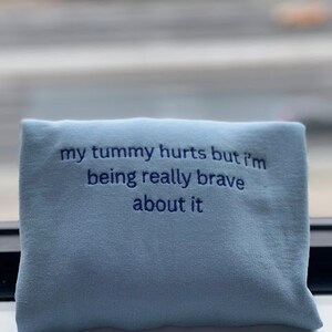 May include: Light blue sweatshirt with the text "my tummy hurts but i'm being really brave about it" embroidered in dark blue.
