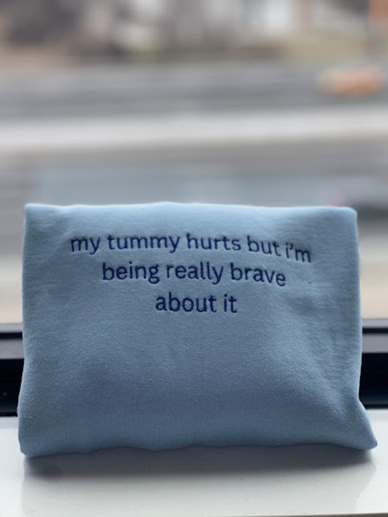 May include: Light blue sweatshirt with the text "my tummy hurts but i'm being really brave about it" embroidered in dark blue.