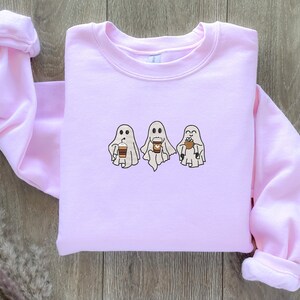 Puede incluir: Una sudadera rosa con un gr&aacute;fico de tres fantasmas sosteniendo tazas de caf&eacute;. Los fantasmas son blancos con ojos y bocas negros. Las tazas de caf&eacute; son marrones con tapas blancas.
