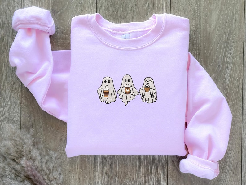 Puede incluir: Una sudadera rosa con un gr&aacute;fico de tres fantasmas sosteniendo tazas de caf&eacute;. Los fantasmas son blancos con ojos y bocas negros. Las tazas de caf&eacute; son marrones con tapas blancas.