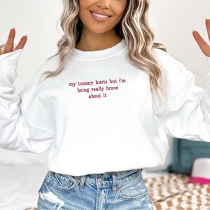 May include: White sweatshirt with the text "my tummy hurts but I'm being really brave about it" in pink.