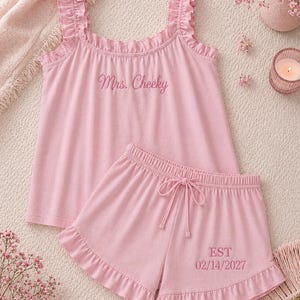 May include: Pink pajama set featuring a ruffled tank top and matching shorts. The tank top has the embroidered text "Mrs. Cheeky", and the shorts have "EST 02/14/2027". The set is made of soft material and is designed for comfort.