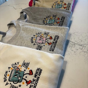 K&ouml;nnte beinhalten: Ein Stapel von f&uuml;nf Sweatshirts in verschiedenen Graut&ouml;nen, Rosa und Beige. Jedes Sweatshirt zeigt eine bunte Illustration eines Buches mit dem Text "A Book A Day" und "Read More Cry Less" &uuml;ber dem Buch. Der Text "Keeps the Mental Breakdown Away" ist unter der Buchillustration gedruckt.