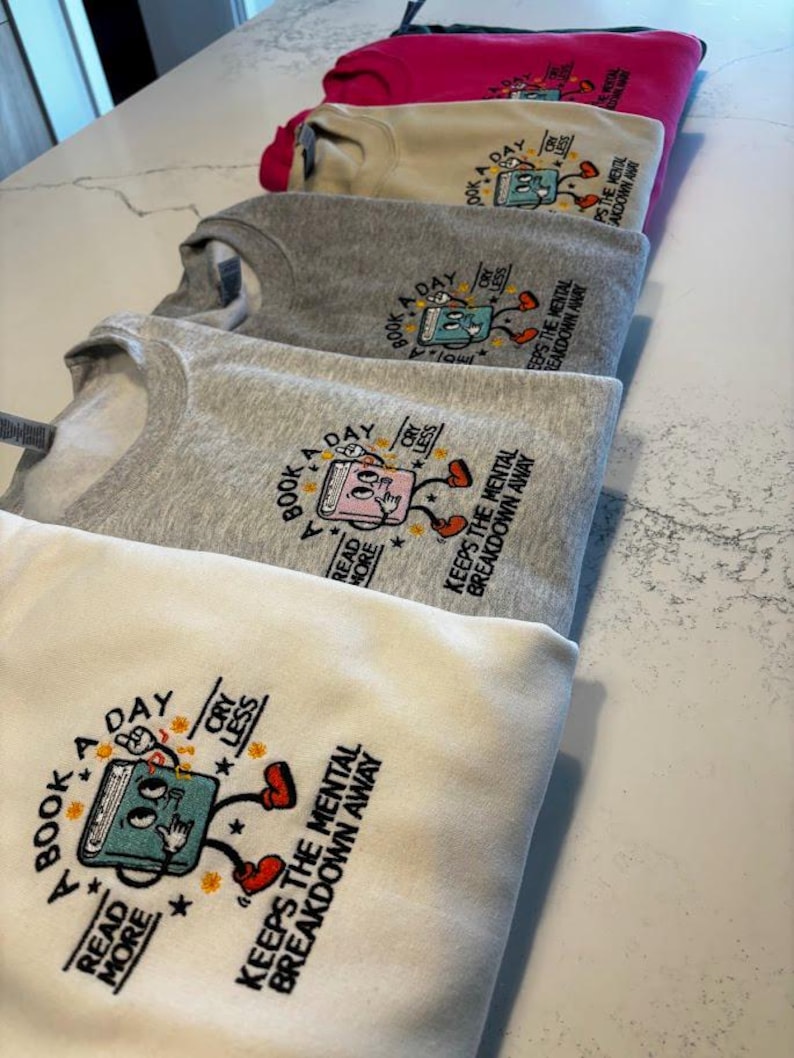 K&ouml;nnte beinhalten: Ein Stapel von f&uuml;nf Sweatshirts in verschiedenen Graut&ouml;nen, Rosa und Beige. Jedes Sweatshirt zeigt eine bunte Illustration eines Buches mit dem Text "A Book A Day" und "Read More Cry Less" &uuml;ber dem Buch. Der Text "Keeps the Mental Breakdown Away" ist unter der Buchillustration gedruckt.