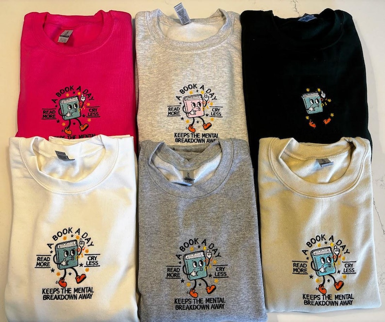 K&ouml;nnte beinhalten: Sechs Rundhals-Sweatshirts in verschiedenen Farben, darunter Pink, Grau, Schwarz, Wei&szlig; und Beige. Jedes Sweatshirt zeigt ein gesticktes Design eines Comic-Buchcharakters mit dem Text "A Book A Day Keeps The Mental Breakdown Away" und "Read More Cry Less".