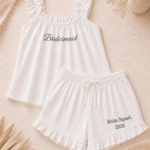 May include: White pajama set with a ruffled top and shorts. The top has the word "Bridesmaid" in green script. The shorts have a drawstring waist and the words "Bride Squad, 2026" in green.