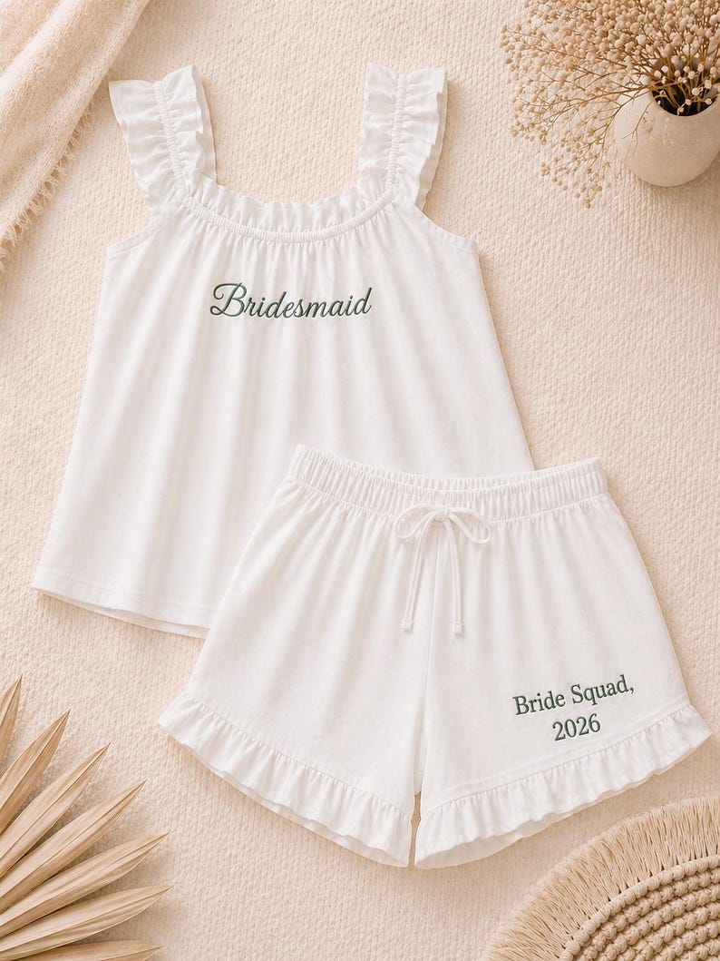 May include: White pajama set with a ruffled top and shorts. The top has the word "Bridesmaid" in green script. The shorts have a drawstring waist and the words "Bride Squad, 2026" in green.