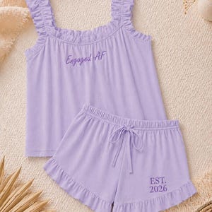 May include: A light purple pajama set featuring a tank top with ruffled straps and the text "Engaged AF". The matching shorts have a drawstring waist and the text "EST. 2026". The set is made of a soft fabric.