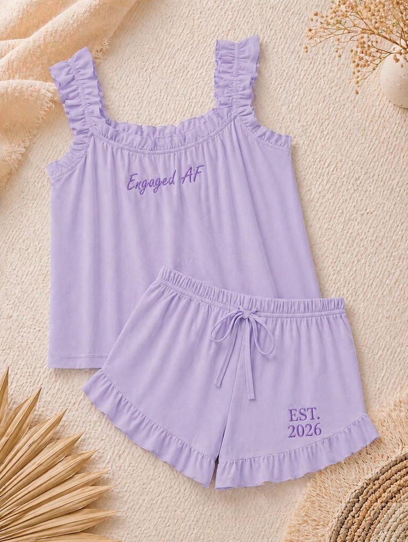May include: A light purple pajama set featuring a tank top with ruffled straps and the text "Engaged AF". The matching shorts have a drawstring waist and the text "EST. 2026". The set is made of a soft fabric.