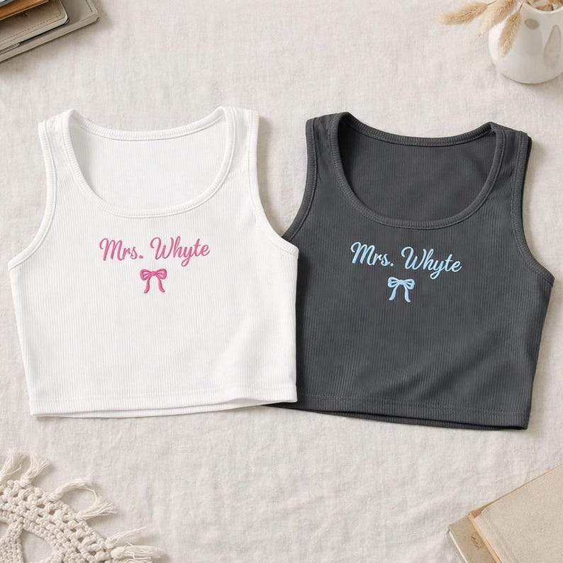 May include: Two ribbed crop tops, one white and one charcoal gray. Each top has the text "Mrs. Whyte" and a bow graphic in a contrasting color. The white top has pink text and bow, while the gray top has blue text and bow.