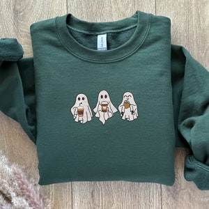 Puede incluir: Una sudadera verde oscuro con un gr&aacute;fico de tres fantasmas sosteniendo tazas de caf&eacute;. Los fantasmas son blancos con ojos y bocas negros.