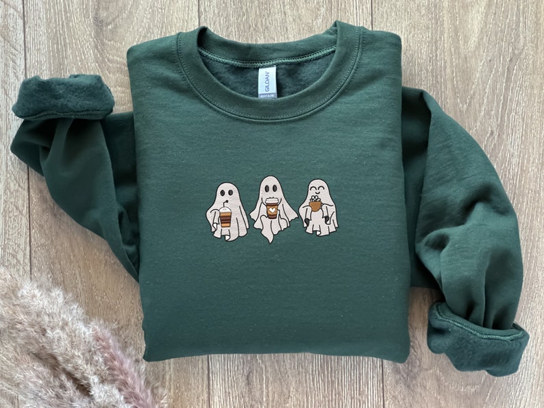 Puede incluir: Una sudadera verde oscuro con un gr&aacute;fico de tres fantasmas sosteniendo tazas de caf&eacute;. Los fantasmas son blancos con ojos y bocas negros.