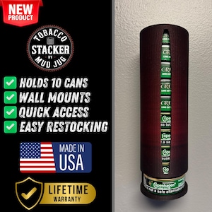 Puede incluir: Un dispensador de latas de tabaco de pared hecho de madera con una mancha de color rojo oscuro. Tiene capacidad para 10 latas y tiene un diseño de acceso rápido. El dispensador está etiquetado como "Tobacco Stacker by Mud Jug" y cuenta con una garantía de por vida. El texto "Made in USA" también es visible.