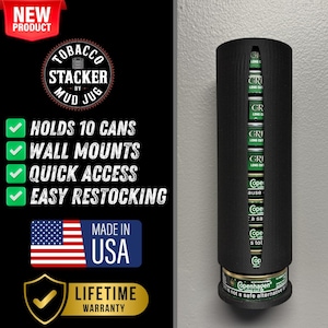 Puede incluir: Un dispensador de latas de tabaco negro de pared con textura de madera. Tiene capacidad para 10 latas y tiene una etiqueta que dice "Copenhagen Long Cut - Not a safe alternative to quitting". El dispensador está etiquetado como "Tobacco Stacker by Mud Jug" y tiene una marca de verificación verde con el texto "Lifetime Warranty".