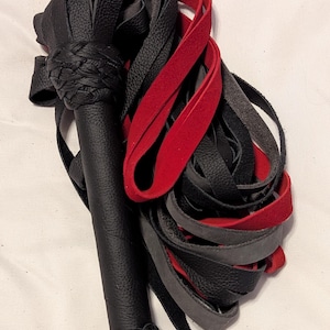 May include: A black leather flogger with a braided handle and a cluster of black, red, and gray leather falls. The handle is cylindrical and has a braided section near the top and bottom.