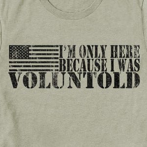 May include: A light olive green t-shirt with a distressed black graphic. The graphic features a stylized American flag and the text "I'M ONLY HERE BECAUSE I WAS VOLUNTOLD."
