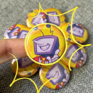 May include: A collection of yellow, round buttons featuring a cartoon television character with a purple screen and a smiling face. The buttons have a yellow border and are arranged on a gray surface.