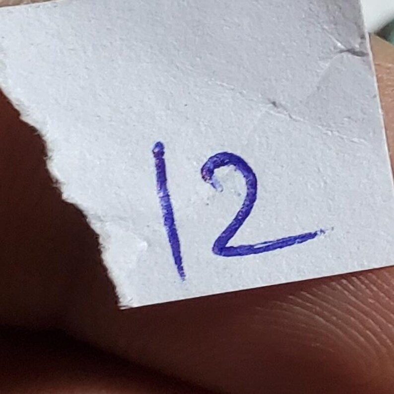 May include: A close-up of a torn piece of white paper with the number 12 written on it in dark blue ink. The number is handwritten and slightly smudged, with the '1' being a straight line and the '2' having a curved top.
