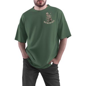 May include: Dark green t-shirt with a graphic of a skeleton on a motorcycle wheel, with the text "WAITING ON PARTS". The shirt has short sleeves and a relaxed fit. The person is wearing dark gray jeans.