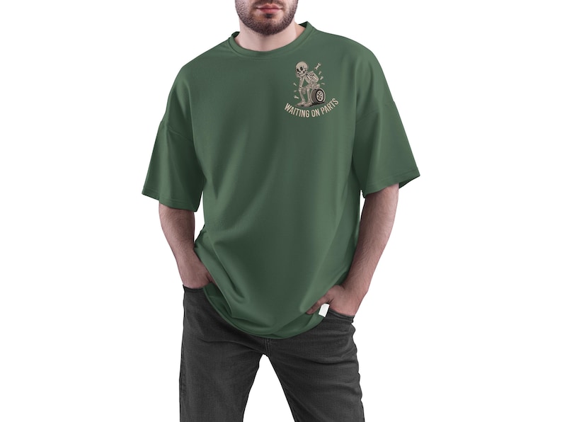 May include: Dark green t-shirt with a graphic of a skeleton on a motorcycle wheel, with the text "WAITING ON PARTS". The shirt has short sleeves and a relaxed fit. The person is wearing dark gray jeans.