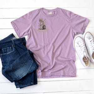 May include: A lilac-colored t-shirt with a graphic of a skeleton pushing a wheel, with the text "Waiting or Dying". The shirt is paired with dark blue jeans and white sneakers with red accents, all arranged on a white wooden surface.