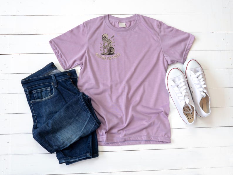 May include: A lilac-colored t-shirt with a graphic of a skeleton pushing a wheel, with the text "Waiting or Dying". The shirt is paired with dark blue jeans and white sneakers with red accents, all arranged on a white wooden surface.