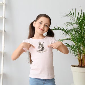 May include: A light pink t-shirt featuring a black and white graphic of a skeleton sitting on a tire, with the text "WAITING ON PARTS". The shirt is being pointed at by a young person.