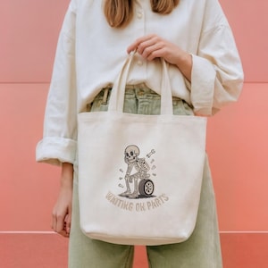 May include: Cream-colored tote bag featuring a cartoon skeleton sitting on a tire, with a wrench and scattered bones. The text "WAITING ON PARTS" is arched below the skeleton. The bag is held against a pink background.