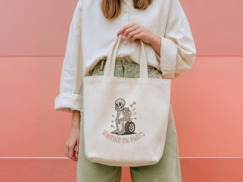 May include: Cream-colored tote bag featuring a cartoon skeleton sitting on a tire, with a wrench and scattered bones. The text "WAITING ON PARTS" is arched below the skeleton. The bag is held against a pink background.