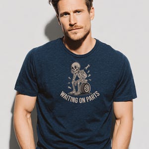 May include: Navy blue t-shirt featuring a graphic of a skeleton sitting on a tire, with wrenches and the text "WAITING ON PARTS". The shirt is made of a soft, comfortable material.