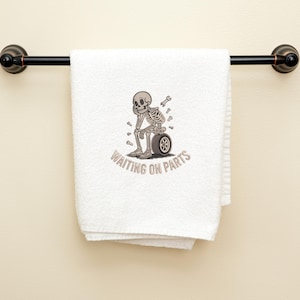 May include: White towel with a black metal bar. The towel features an embroidered skeleton sitting on a tire, with the text "WAITING ON PARTS" in a curved font. The skeleton has a wrench and other tools nearby.