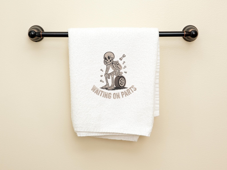 May include: White towel with a black metal bar. The towel features an embroidered skeleton sitting on a tire, with the text "WAITING ON PARTS" in a curved font. The skeleton has a wrench and other tools nearby.