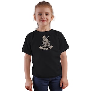 May include: Black t-shirt featuring a skeleton design with the text "WAITING ON PARTS". The graphic shows a skeleton sitting on a tire, with a thoughtful expression. The shirt is worn by a young person.