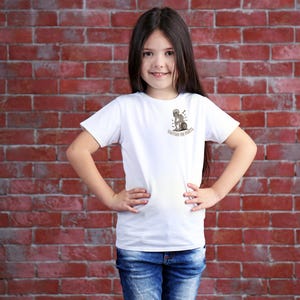 May include: White t-shirt with a small graphic of a skeleton riding a motorcycle and the text "Riding the Parts". The shirt is worn by a person in blue jeans, standing in front of a red brick wall.