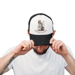 May include: A black and white trucker hat with a graphic of a skeleton sitting on a tire, with the text "WAITING ON PARTS". The hat has a black brim and mesh back.
