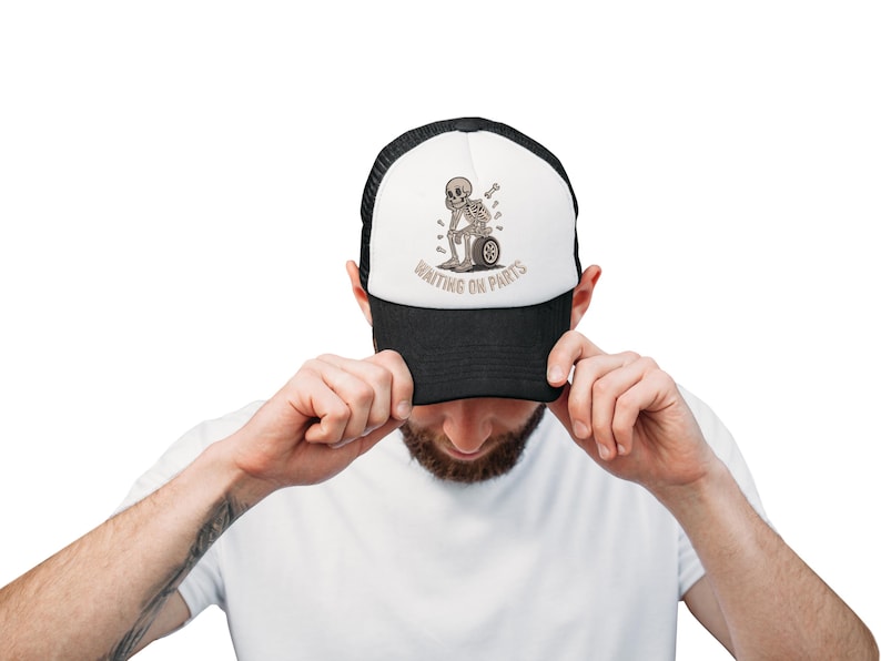 May include: A black and white trucker hat with a graphic of a skeleton sitting on a tire, with the text "WAITING ON PARTS". The hat has a black brim and mesh back.