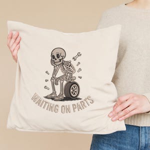 May include: Beige throw pillow with a distressed graphic of a skeleton sitting on a tire, surrounded by wrenches. The text "WAITING ON PARTS" is printed below the skeleton in a matching font.