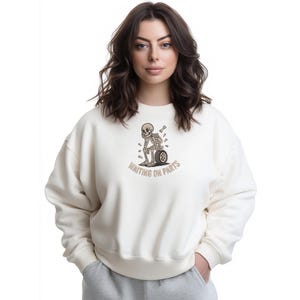 May include: Cream-colored sweatshirt with a graphic of a skeleton leaning against a tire, with the text "WAITING ON PARTS" in brown. The sweatshirt has a relaxed fit and long sleeves. The model is wearing gray sweatpants.