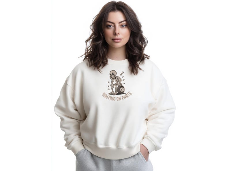 May include: Cream-colored sweatshirt with a graphic of a skeleton leaning against a tire, with the text "WAITING ON PARTS" in brown. The sweatshirt has a relaxed fit and long sleeves. The model is wearing gray sweatpants.