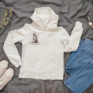 May include: A light gray hoodie with a front pocket and a graphic of a skeleton on a tire, with the text "Waiting on Parts." The hoodie is paired with blue jeans, white sneakers, and a watch.