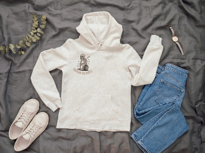 May include: A light gray hoodie with a front pocket and a graphic of a skeleton on a tire, with the text "Waiting on Parts." The hoodie is paired with blue jeans, white sneakers, and a watch.