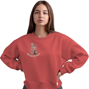 May include: Coral-colored sweatshirt featuring a graphic of a skeleton sitting on a motorcycle wheel, with the text "WAITING ON PARTS." The crew neck sweatshirt is a casual style.
